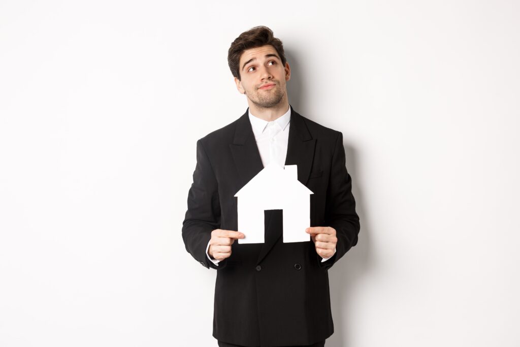 A man in a business suit holding a paper cutout of a house, looking up thoughtfully, against a plain white background.