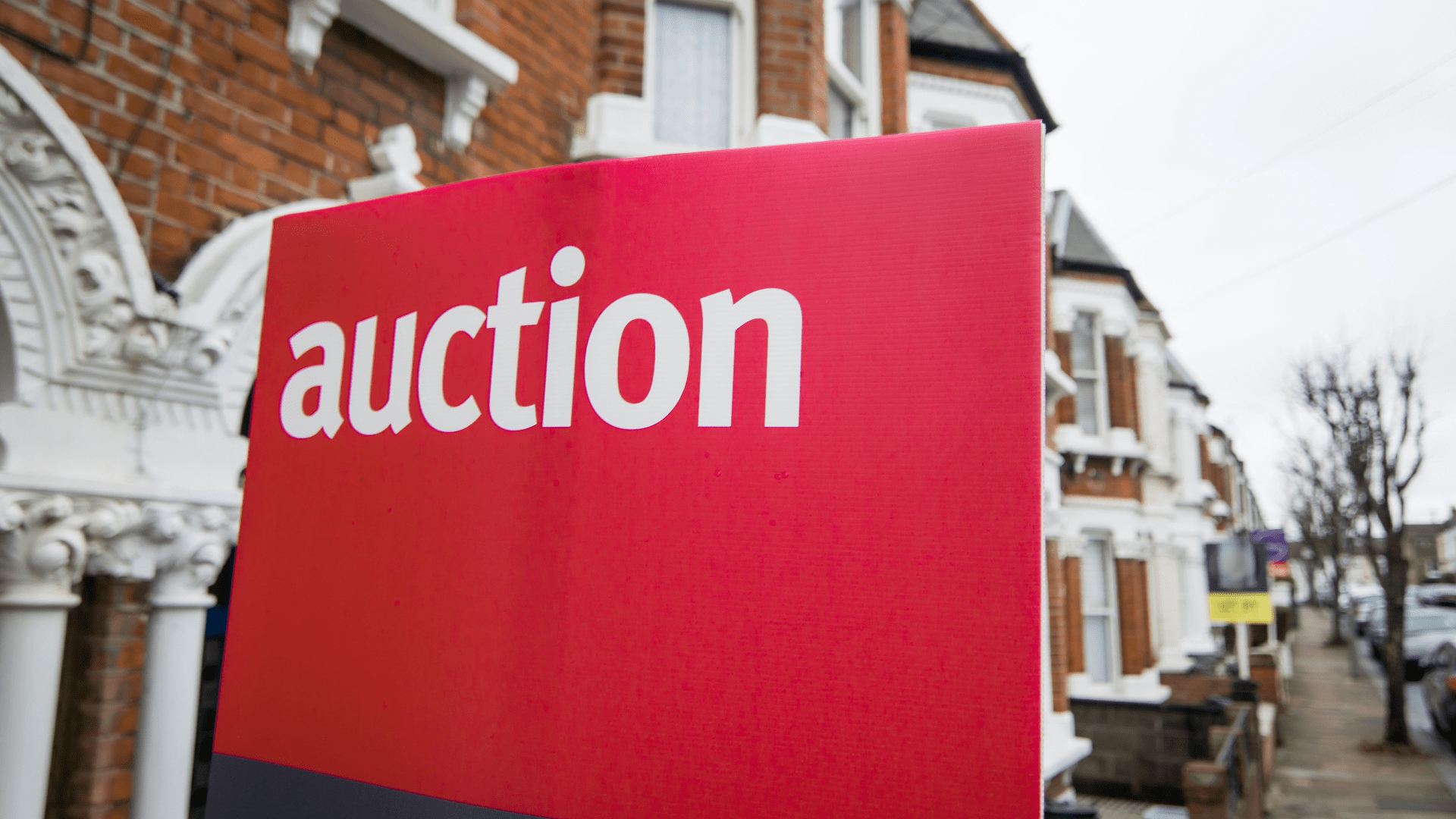 A prominent red "auction" sign in the foreground, with a blurred background of a residential street lined with Victorian-style houses.