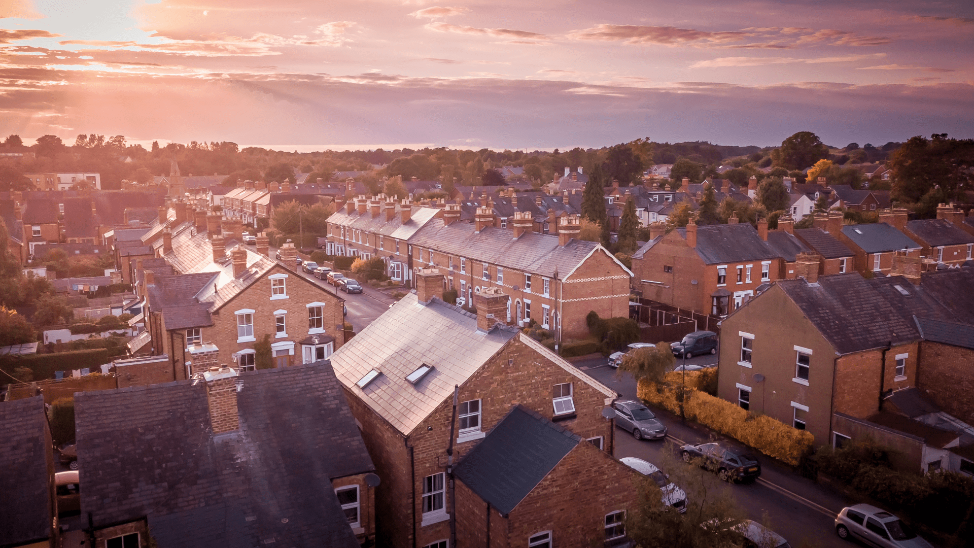 Aerial view of a residential area at sunset, featuring rows of brick houses with slanted roofs and chimneys, bathed in a soft golden light.