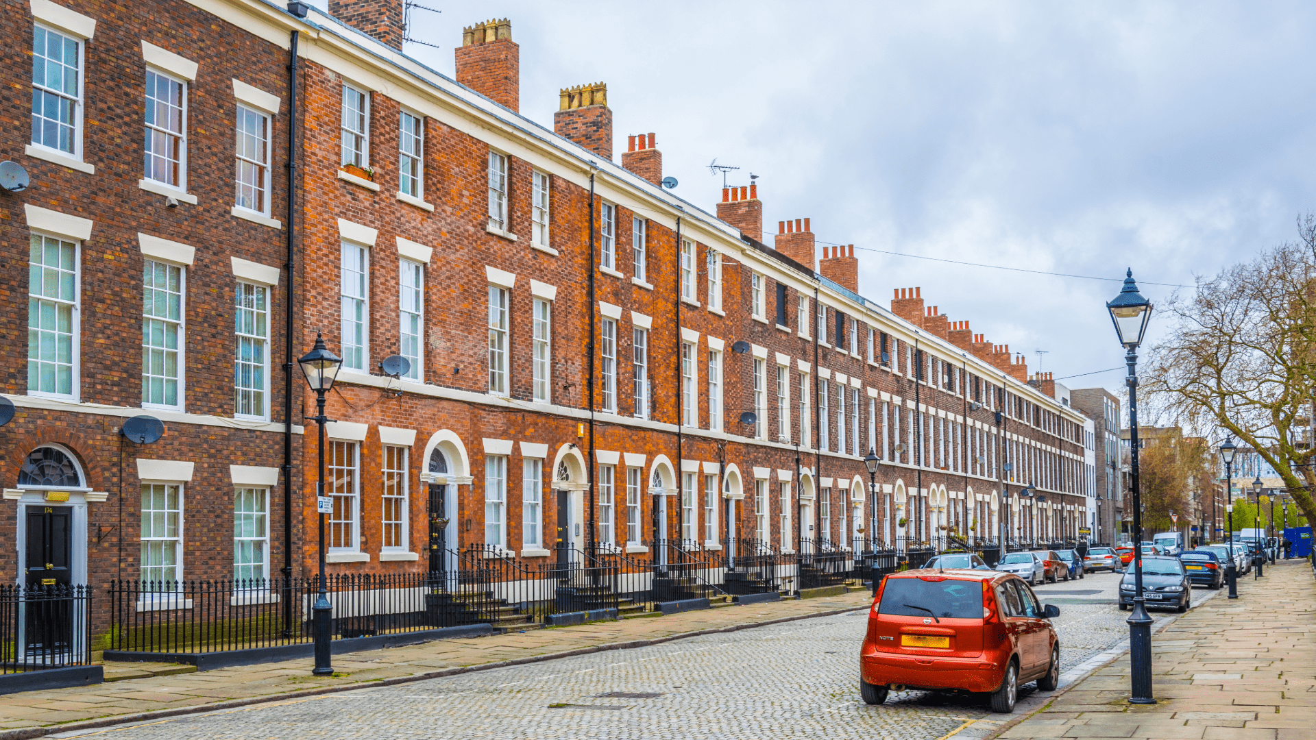 A traditional Georgian terrace on a cobbled street in the UK, with rows of red brick houses, white framed windows, and black doors, under an overcast sky.