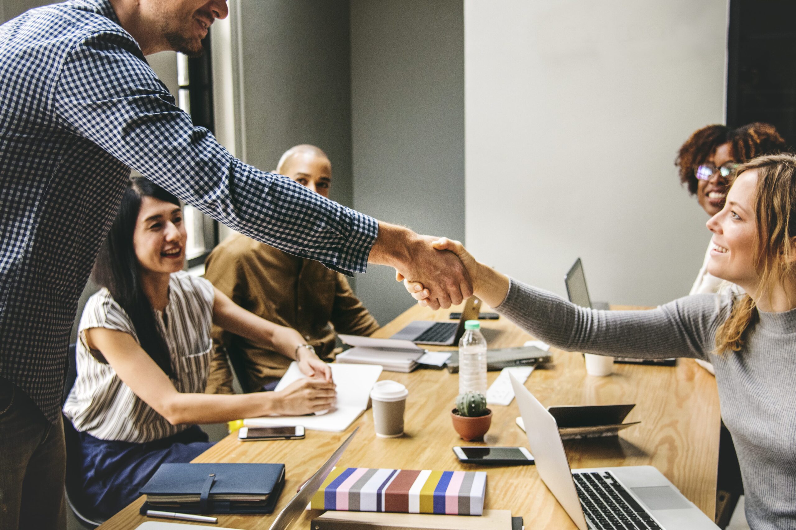 Business meeting with people shaking hands across a table, laptops and notebooks in front of them.