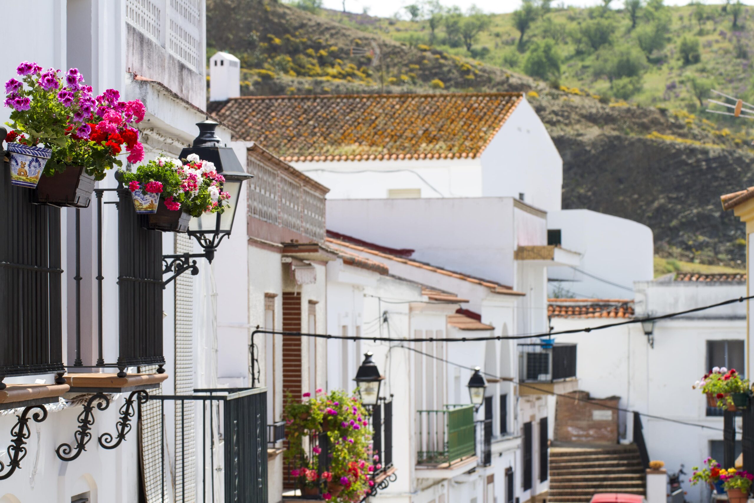 Charming Spanish village street with white houses, balconies adorned with colorful flowers, and a mountainous backdrop.