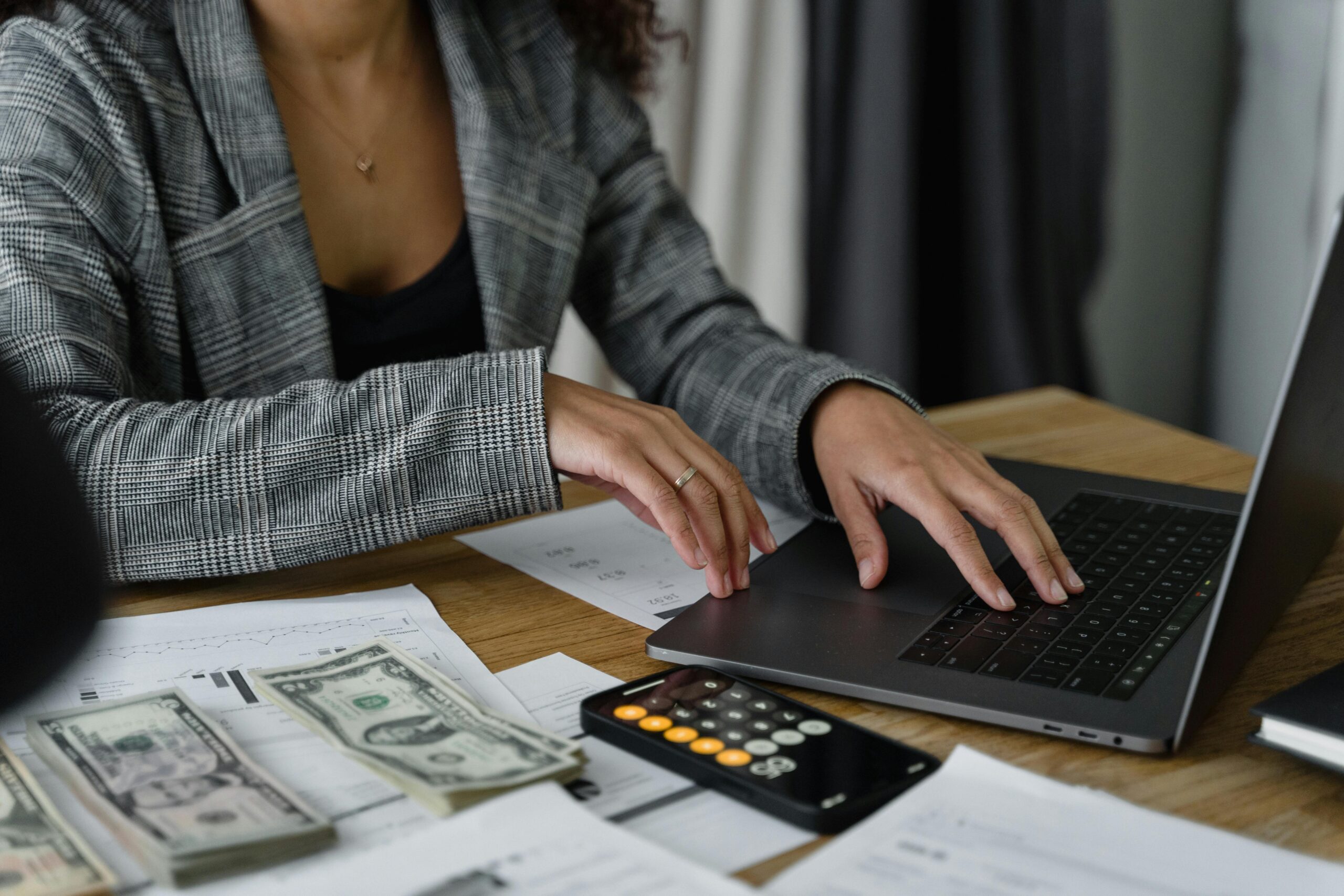 Person working on a laptop with financial documents, cash, and a calculator on the desk.